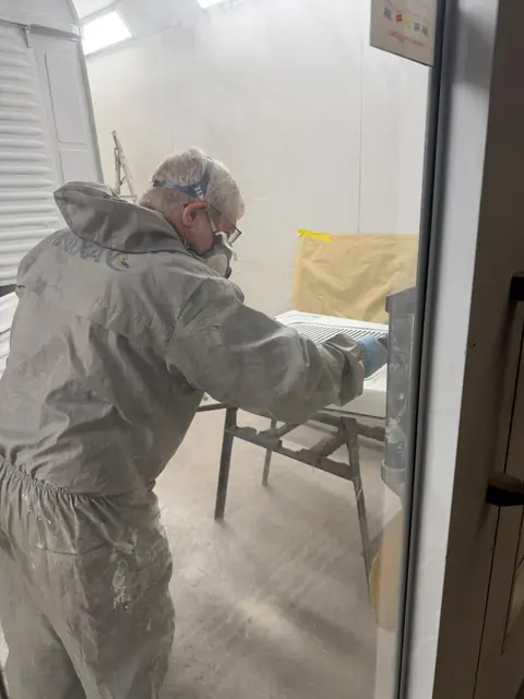 Mark in overalls and a respirator preparing a white panel inside the spray booth